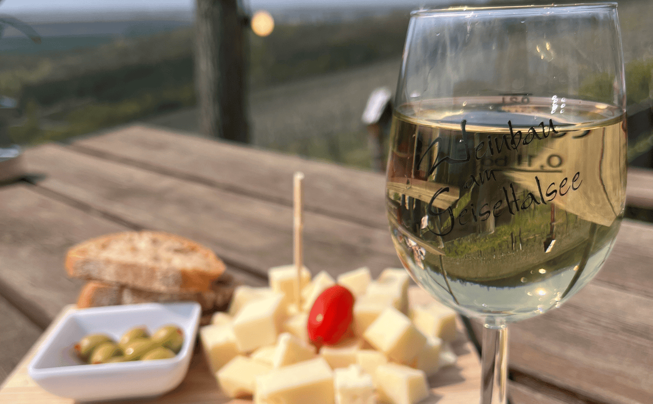 DSW-Mitgliedervertreterfahrt-Geiseltalsee-BB-05 Glas Weißwein mit Blick auf die Weinregion Naumburg, Käse, Oliven und Baguette auf einem Holztisch im Freien.