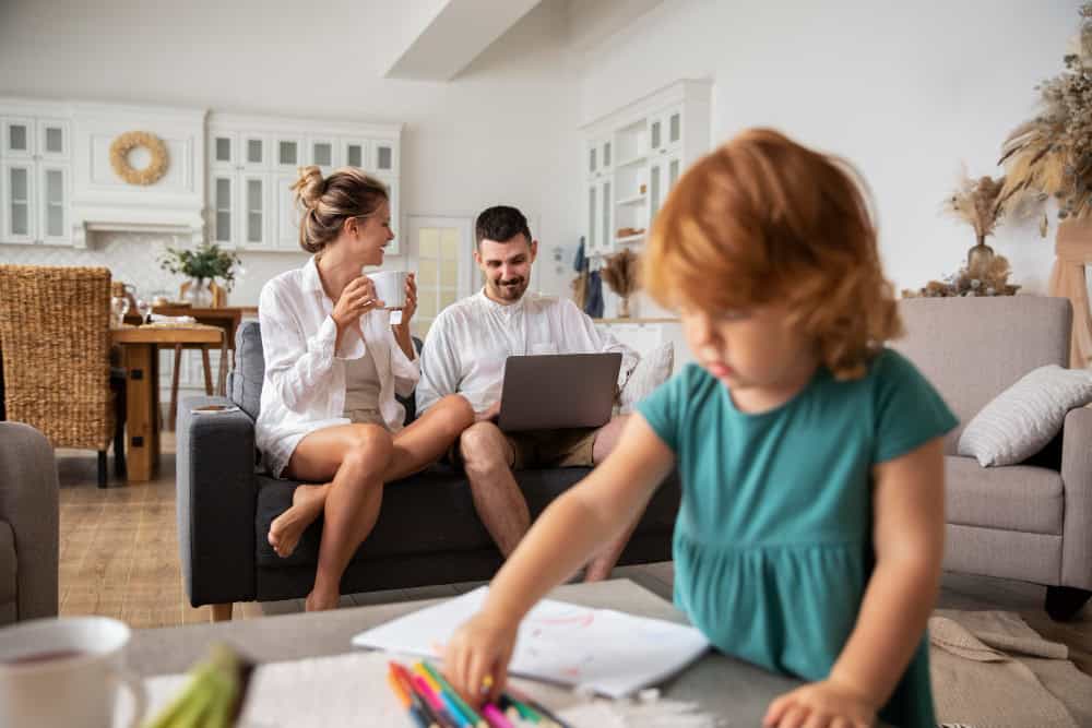 Eltern beantragen Kindergeld am Laptop auf dem Sofa. Kind spielt im Vordergrund.